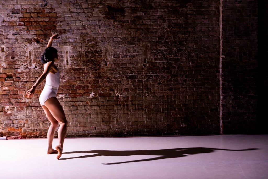 A person in a tight white suit in front of a brick wall, harsh light from the left casting a thin sharp black shadow of the person. She has the left arm raised and the right one swinging down to the right.